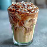 Iced Vanilla Bean Frappuccino with Coconut Whipped Cream in a clear glass, topped with fluffy coconut cream and a sprinkle of vanilla bean powder.