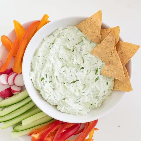 A close-up of vibrant Green Goddess Salad Dip in a white bowl, garnished with fresh parsley and tarragon leaves, surrounded by crunchy tortilla chips and colorful veggie sticks.