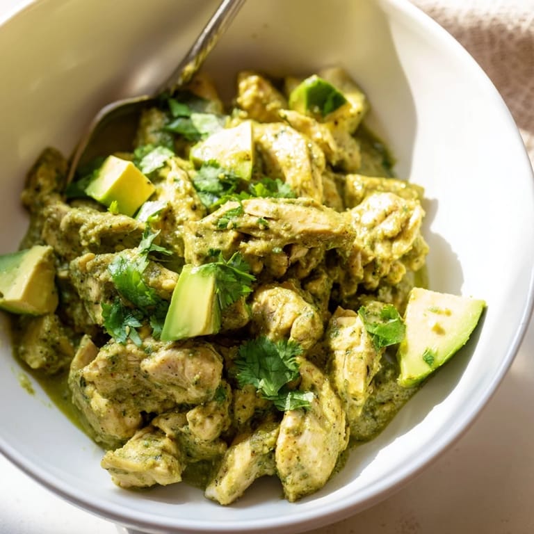A close-up of flavorful chicken chili verde topped with creamy avocado slices, fresh cilantro, and a lime wedge beside warm corn tortillas.